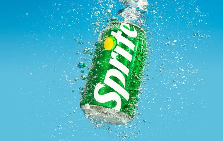 A silver Sprite can exploding through a massive splash of clear water against a blue background.